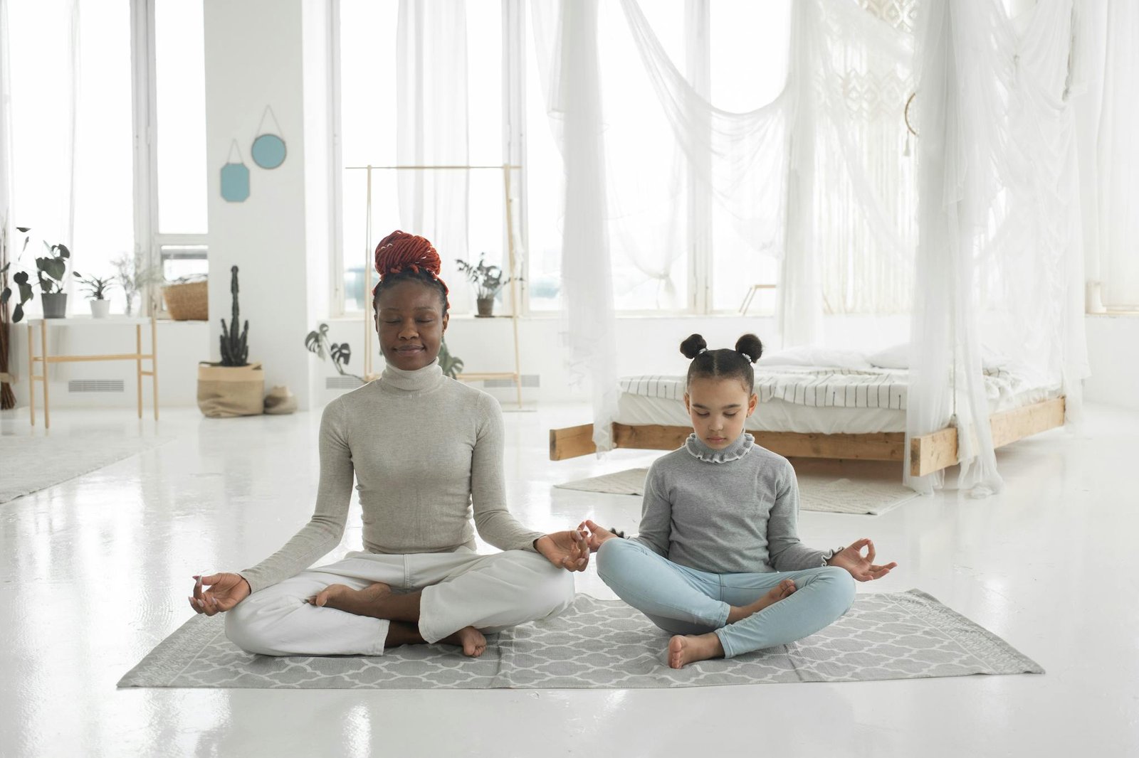 Full body of concentrated young barefooted black woman with cute little daughter sitting together on floor in Padmasana pose and mudra hands and meditating with closed eyes in light bedroom