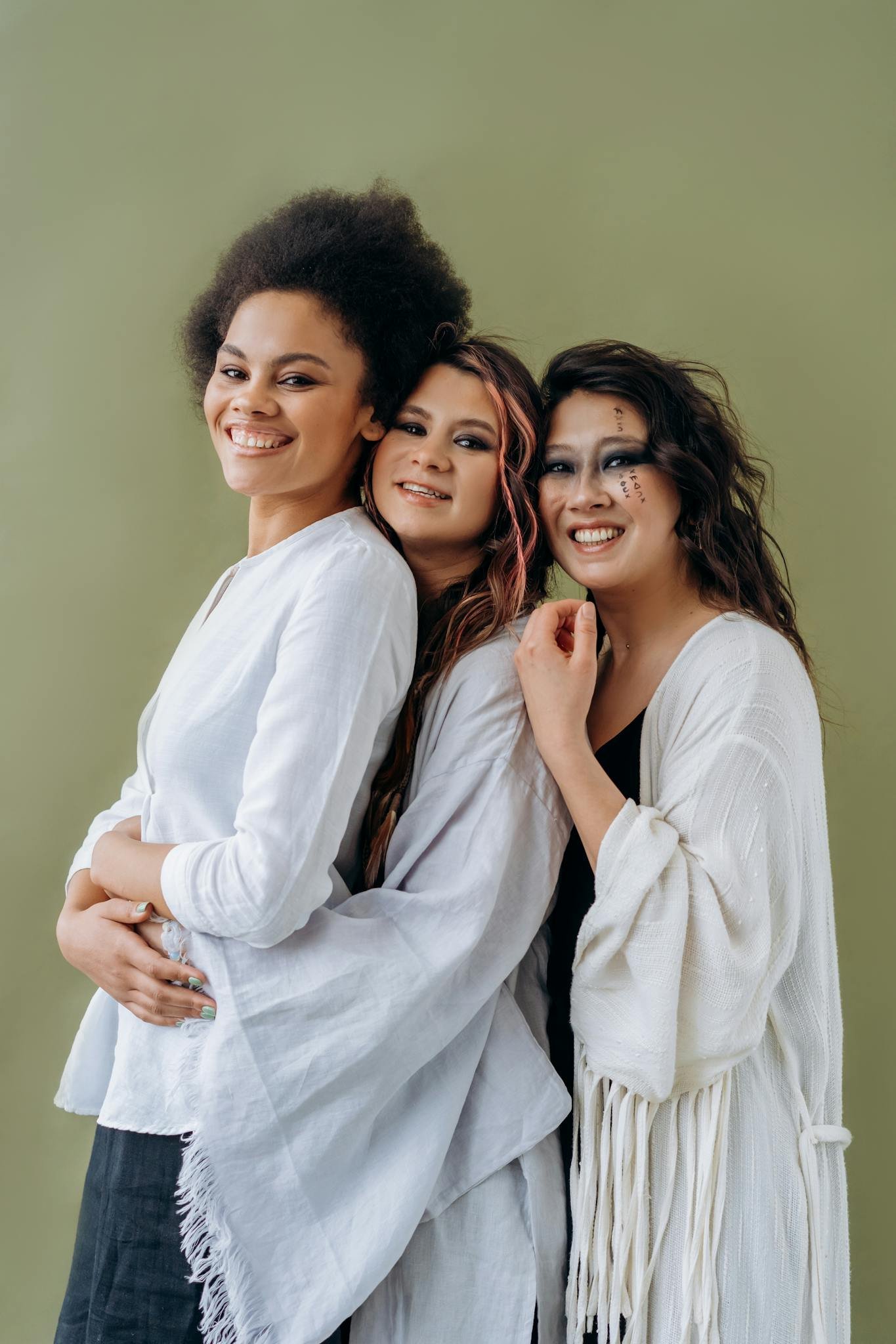 Three multicultural women in white attire stand happily embracing against a green backdrop.